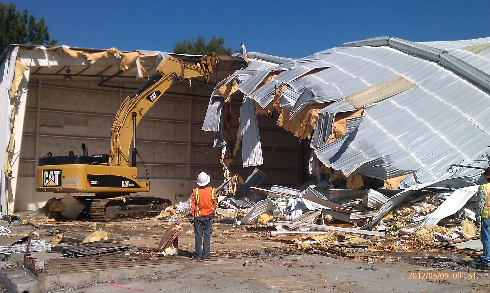 Excavator tearing down a commercial building's metal roof under a clear blue sky